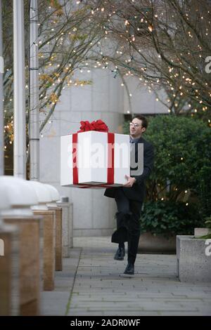 Un giovane uomo che porta un grande regalo bianco avvolto con un fiocco rosso passeggiando attraverso un cortile Foto Stock