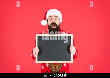 Incredibile. Annuncio d'inverno. Evento invernale. Vacanze invernali. Man tenere lavagna vuota copia spazio. Guy babbo natale sfondo rosso. Santa Claus hat con la pubblicità. Lavagna per informazioni. Foto Stock
