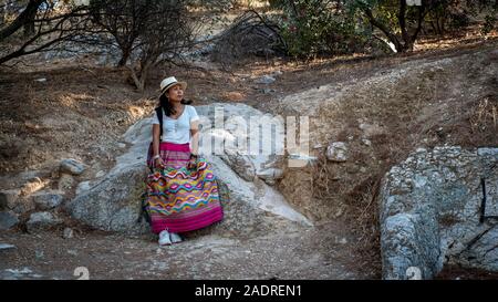 Donna asiatica in t-shirt bianco ad Atene nel cappello bianco sneakers e sarong appoggiata contro la roccia in Agora Foto Stock