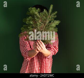 Una donna in una maglietta rossa è in piedi contro un muro verde, tenendo un albero di Natale ramo nelle sue mani e che copre il volto. Concetto di vacanza. Minimali Foto Stock