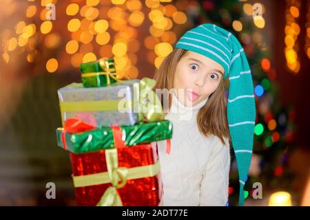 Un funny girl in un gnome hat stand con molti doni nelle sue mani contro lo sfondo di brillanti luci delle vacanze. Natale e Anno Nuovo. Foto Stock