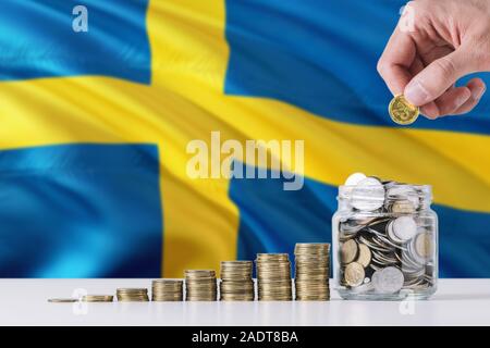 Business man holding monete mettendo in vetro, Svezia bandiera sventola in background. Finanza e la concezione di business. Risparmio di denaro. Foto Stock