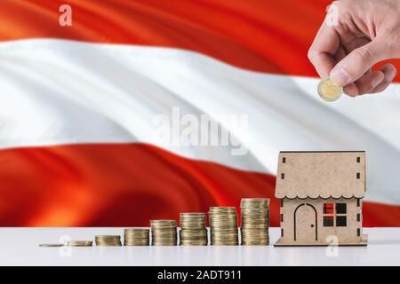 Uomo con monete mettendo in casa di legno salvadanaio, Austria bandiera sventola in background. Risparmio di denaro per i mutui ipotecari. Foto Stock