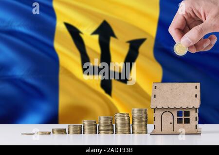 Uomo con monete mettendo in casa di legno salvadanaio, Barbados bandiera sventola in background. Risparmio di denaro per i mutui ipotecari. Foto Stock