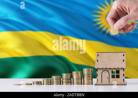 Uomo con monete mettendo in casa di legno salvadanaio, Ruanda bandiera sventola in background. Risparmio di denaro per i mutui ipotecari. Foto Stock