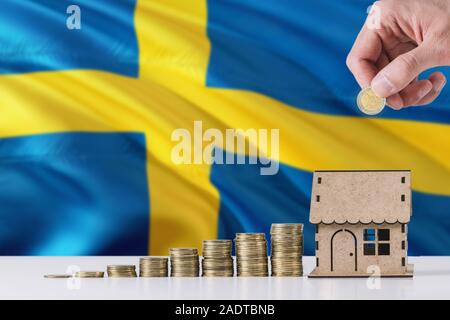 Uomo con monete mettendo in casa di legno salvadanaio, Svezia bandiera sventola in background. Risparmio di denaro per i mutui ipotecari. Foto Stock