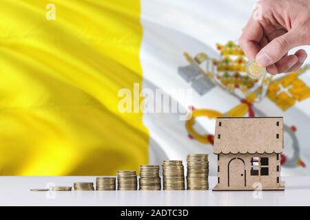 Uomo con monete mettendo in casa di legno salvadanaio, Città del Vaticano bandiera sventola in background. Risparmio di denaro per i mutui ipotecari. Foto Stock