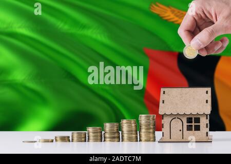 Uomo con monete mettendo in casa di legno salvadanaio, Zambia bandiera sventola in background. Risparmio di denaro per i mutui ipotecari. Foto Stock