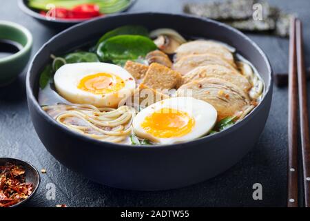 Asian noodle soup, ramen con pollo, tofu, verdure e uova in ciotola nero. Sfondo di ardesia. Close up Foto Stock