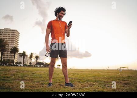 Corpo pieno colpo di un felice athletic giovane azienda di acqua in bottiglia in mano ascoltando musica in cuffia utilizzando smart phone mentre in piedi nel parco Foto Stock