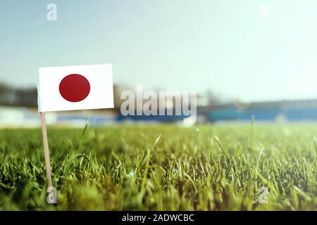 Stick miniaturizzato bandiera del Giappone su erba verde, vicino campo di sole. Lo sfondo dello stadio, copia spazio per il testo. Foto Stock