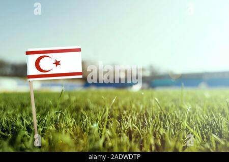 Stick miniaturizzato di Cipro Nord bandiera su erba verde, vicino campo di sole. Lo sfondo dello stadio, copia spazio per il testo. Foto Stock