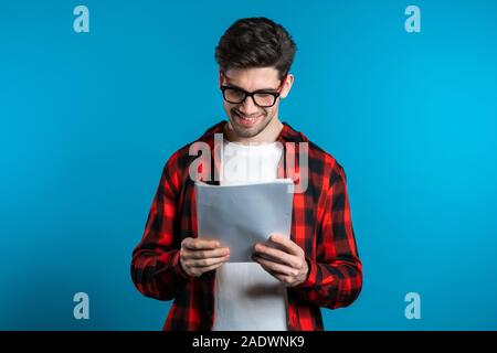 Giovane uomo bello controlla i documenti, bollette, relazione. Imprenditore isolato su sfondo blu. È soddisfatto con il lavoro del personale aziendale. Foto Stock