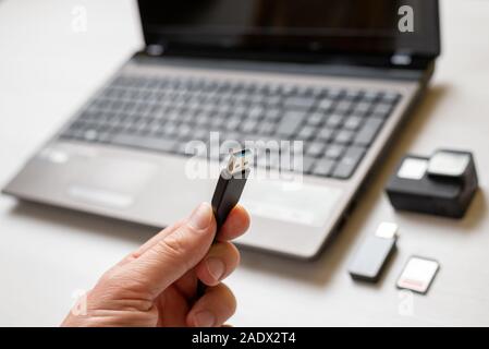 Cavo USB in mano di close-up. Azione la fotocamera e il computer portatile in background. Nozione di trasferimento di foto e video. Foto Stock