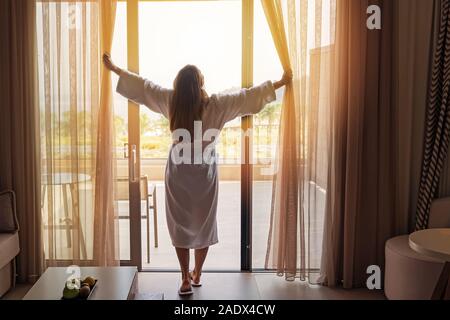 Giovane donna indossare accappatoio bianco apertura tende in camera di questo hotel di lusso Foto Stock
