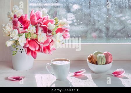 Tazza di caffè sul bordo della finestra e il sole dopo la pioggia. Mazzo di fiori di primavera: tulipani rosa e bianco fresia. Rosa e verde di amaretti, gustosi dolci Foto Stock