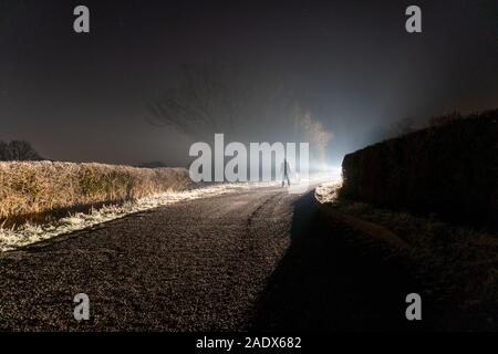 Un uomo stagliano contro una misteriosa luce brillante, incandescente su un vicolo del paese, in una nebbiosa inverni di notte Foto Stock