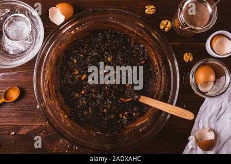 Spessore di pasta al cioccolato con dadi per la deliziosa torta brownie in una grande ciotola con cucchiaio in legno scuro tabella accanto a ingredienti e utensili da cucina, parte superiore Foto Stock