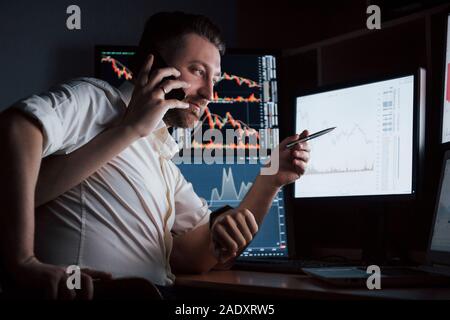 Uomo con molte mani. Barbuto ragazzo in maglia bianca lavora in ufficio con più schermi di computer in tabelle di indice Foto Stock