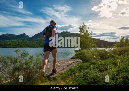 Runner maschio in esecuzione su di un sentiero di montagna. Treni atleta dal lago di montagna. Vista posteriore. Uomo in maglia blu e pantaloncini neri all'aperto di formazione Foto Stock