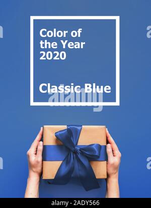 Mani femminili tenere confezione regalo su sfondo blu con le parole il colore dell'anno 2020 Blu Classico. Confezione regalo in craft incarto di carta blu con nastro di raso. Verticale. Foto Stock
