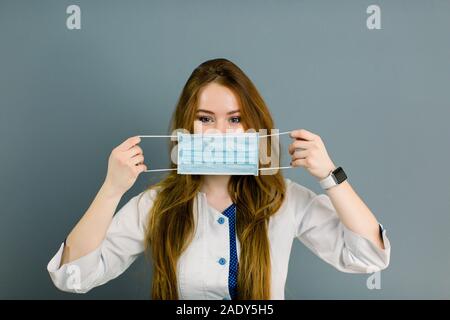 Ritratto di giovane donna medico azienda maschera medico guardando la telecamera isolato su grigio Foto Stock