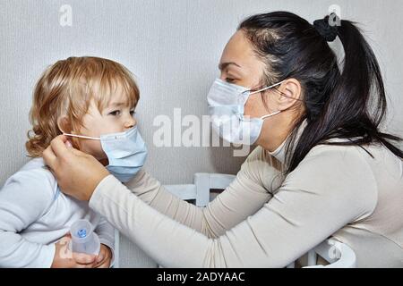 Madre aiuta la sua piccola figlia con malattia cronica di mettere medical maschera sul volto della ragazza. Maschera di protezione da infezioni durante la malattia. Foto Stock