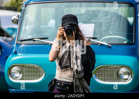 Messa a fuoco selettiva su giovani caucasici donna fotografo tiene la fotocamera reflex digitale nella parte anteriore del vecchio blu van, indossare un cappello nero sciarpa, durante un'auto d'epoca mostrano Foto Stock