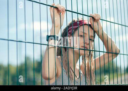 Ritratto di donna calma con dreadlocks guardando a voi attraverso la catena collegamento recinto Foto Stock