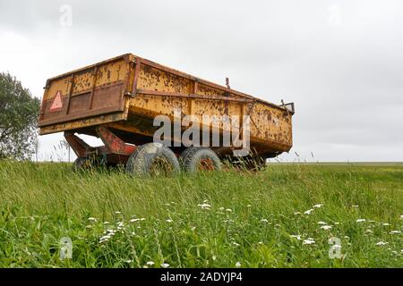 Abbandonato il vecchio giallo arrugginito trattore agricolo carro nel verde paesaggio sotto un grigio cielo nuvoloso con spazio di copia Foto Stock