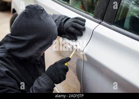 Ladro di auto in azione, un uomo in una cappa di aspirazione si rompe un cacciavite porta auto. L'uomo vestito di nero cercando di penetrare nella macchina. Foto Stock