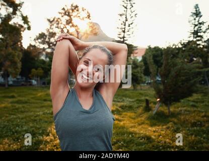 Ritratto di un sorridente sportivo giovane donna stretching le braccia nel parco Foto Stock