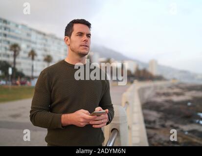 Ritratto di un pensieroso bel giovane uomo in piedi su una strada di città azienda telefono cellulare in mano alla ricerca di distanza Foto Stock