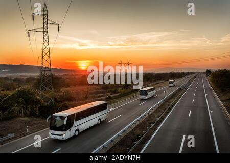 Roulotte o un convoglio di quattro autobus di linea che viaggiano in un paese autostrada sotto incredibile arancione tramonto Cielo. Autostrada il trasporto con autobus di colore bianco Foto Stock