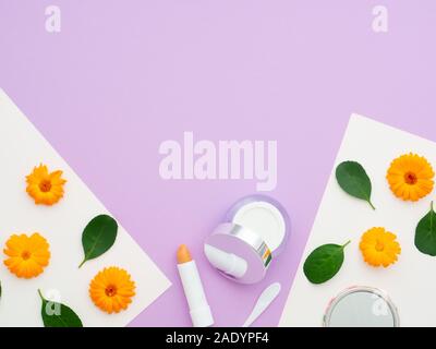 Freschi di fiori e foglie verdi su white paper e cosmetici naturali, crema e balsamo per labbra isolate su uno sfondo viola. Cornice fatta di fiori d'arancio Foto Stock