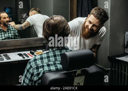 Dai capelli scuri uomo seduto nel barbiere e il suo barbiere sorridente Foto Stock