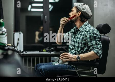 Calma uomo seduto nella sedia da barbiere e spazzolatura della sua barba Foto Stock