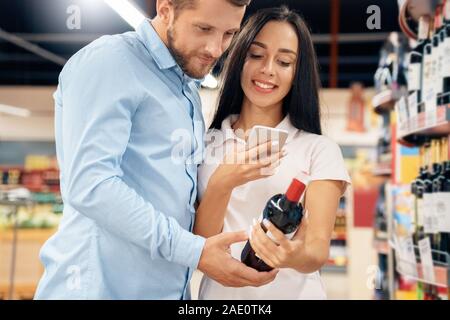 Spesa giornaliera. Matura al supermercato insieme al dipartimento di alcool di vino scegliendo la scansione del codice QR sullo smartphone cerchi informazioni gioiosa close-up Foto Stock
