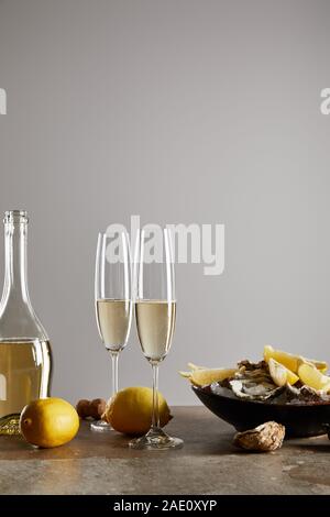 Bicchieri di champagne con vino frizzante vicino a bottiglia, ostriche e limoni nel recipiente isolato su grigio Foto Stock