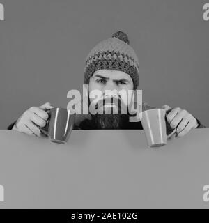 Caffeinated bevande idea. Hipster con la barba e tricky faccia ha il caffè. In autunno il caffè e il barista home concept. Uomo in caldo hat detiene il marrone e verde sulla tazza verde e lo sfondo arancione, spazio di copia Foto Stock