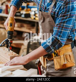 Vista ritagliata del falegname in grembiule azienda trapano martello vicino a tavola in legno Foto Stock