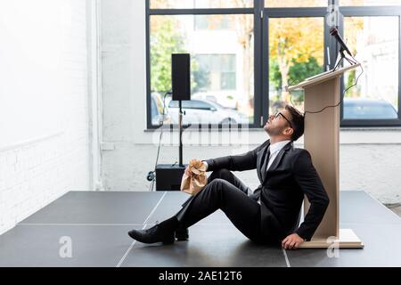 Vista laterale della paura di imprenditore nel soddisfare azienda sacchetto di carta e seduto sul pavimento durante la conferenza Foto Stock