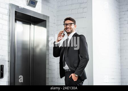 Imprenditore sorridente in formale parlare di usura sullo smartphone vicino all'ascensore Foto Stock