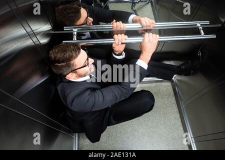 Angolo di alta vista di paura imprenditore della tuta con claustrofobia seduto sul pavimento in ascensore Foto Stock