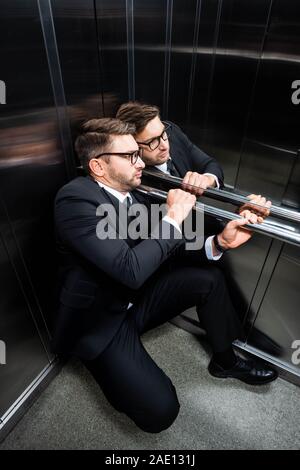 Angolo di alta vista di paura imprenditore della tuta con claustrofobia seduto sul pavimento in ascensore Foto Stock