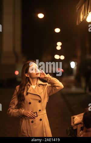 Pensieroso giovane donna in trench coat che guarda lontano e toccando i capelli mentre in piedi sulla strada di sera Foto Stock