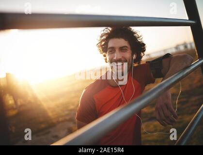 Ritratto di un sorridente giovane atleta maschio con gli auricolari nelle orecchie appoggiato sulle barre orizzontali in appoggio al termine della corsa Foto Stock