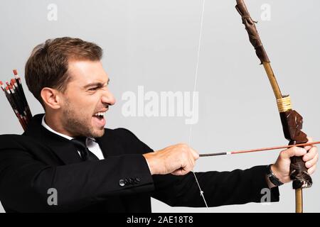 Vista laterale di angry businessman in tuta holding bow e riprese con la freccia isolato su grigio Foto Stock