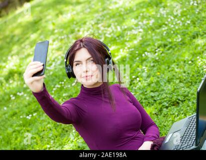 Bella donna giovane studente nel parco utilizzando computer portatile ascolto di musica con cuffie prendere un selfie tramite telefono. Foto Stock