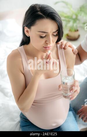 Donna incinta con dolore prendendo la pillola con acqua in camera da letto con il marito vicino Foto Stock